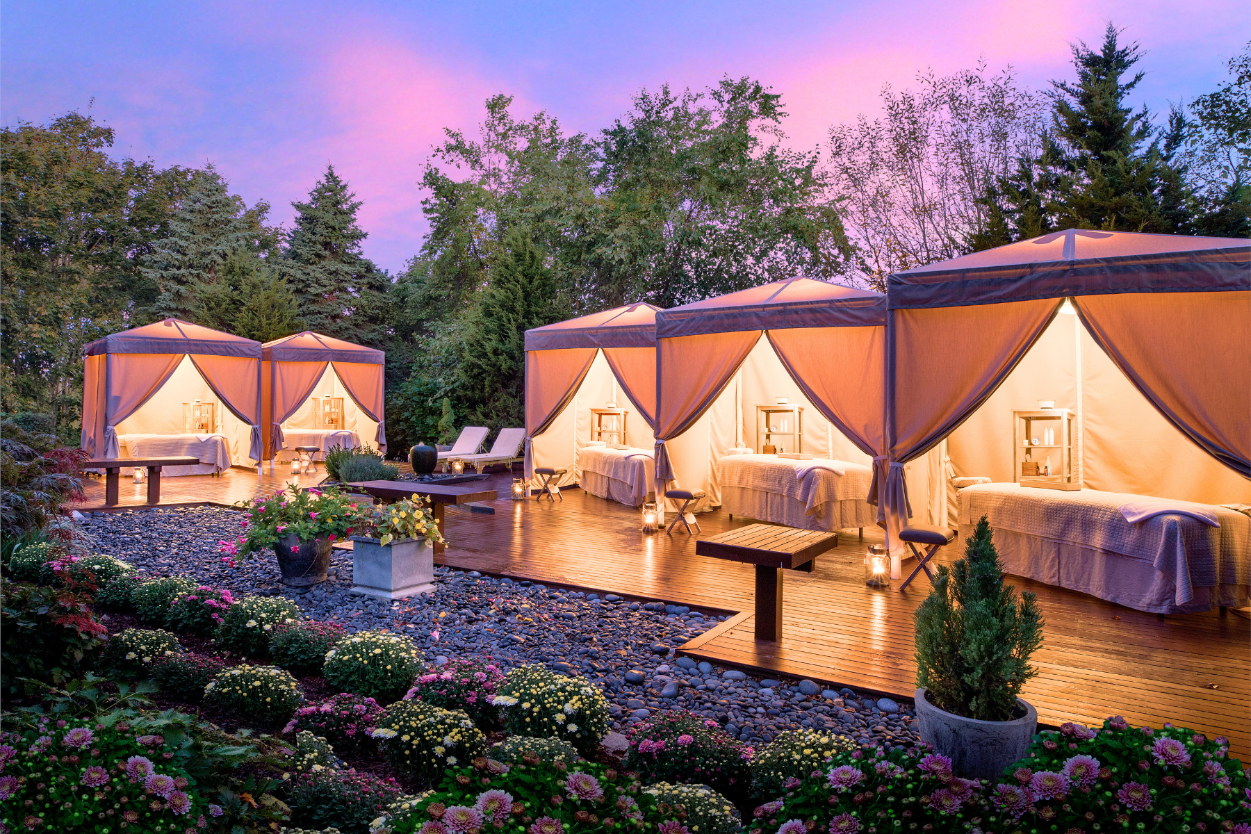 Outdoor spa cabanas with massage tables, surrounded by lush greenery and flowers under a twilight sky.