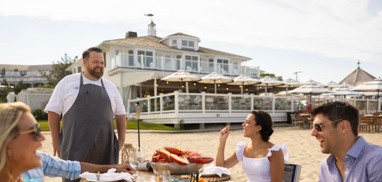 People dining outdoors by the beach, with a chef standing nearby. A large building and umbrellas in the background.