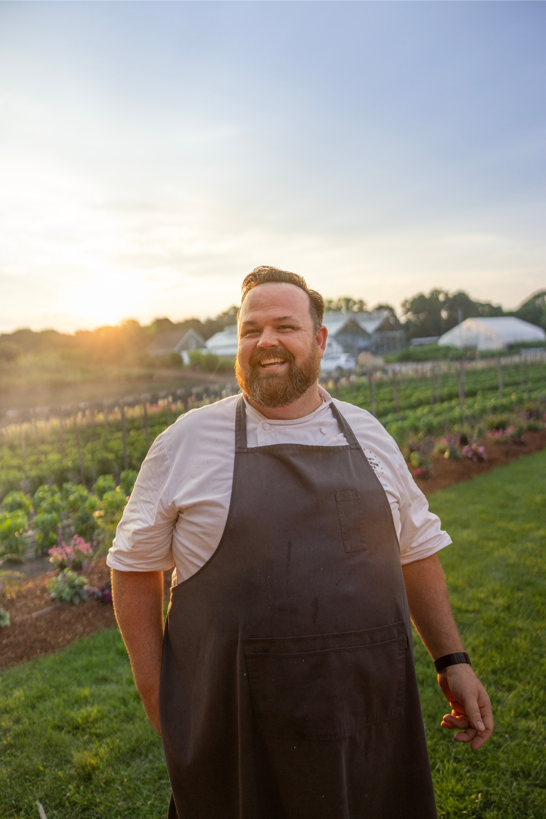 A smiling person in a chef's apron stands in a garden at sunset, with green fields and buildings in the background.