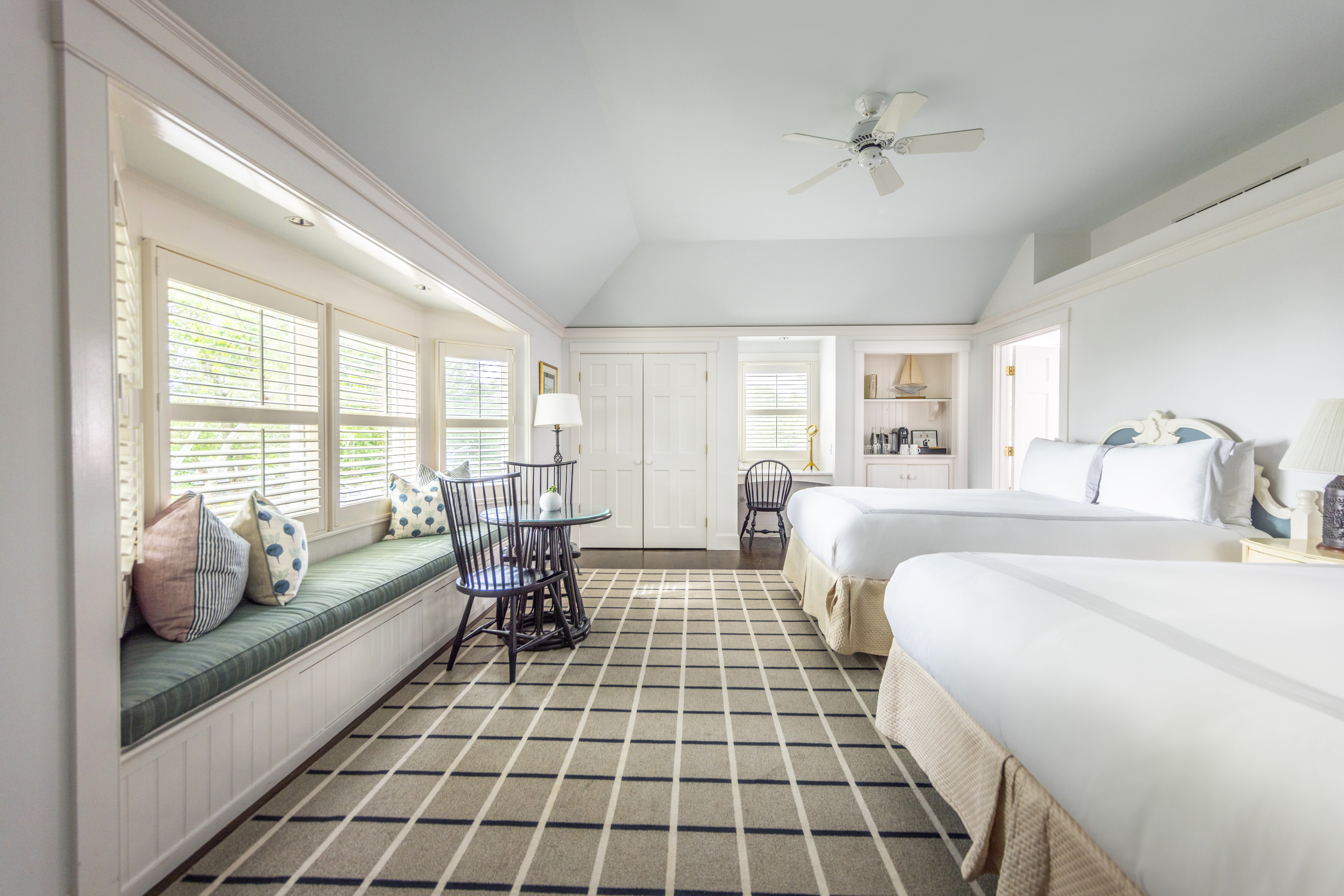 A bright, cozy hotel room with two beds, a window seat, patterned carpet, wooden chairs, and a table, featuring white and pastel tones.