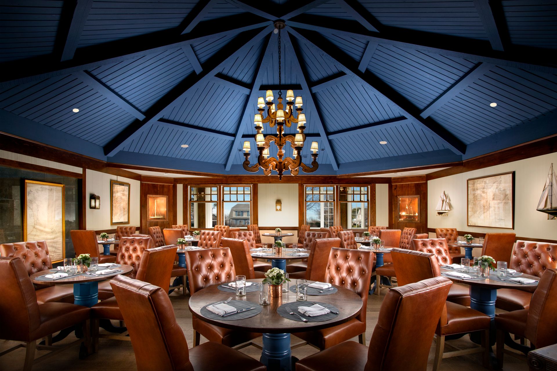 A stylish restaurant interior with round tables, leather chairs, nautical decor, and a blue vaulted ceiling with a chandelier.