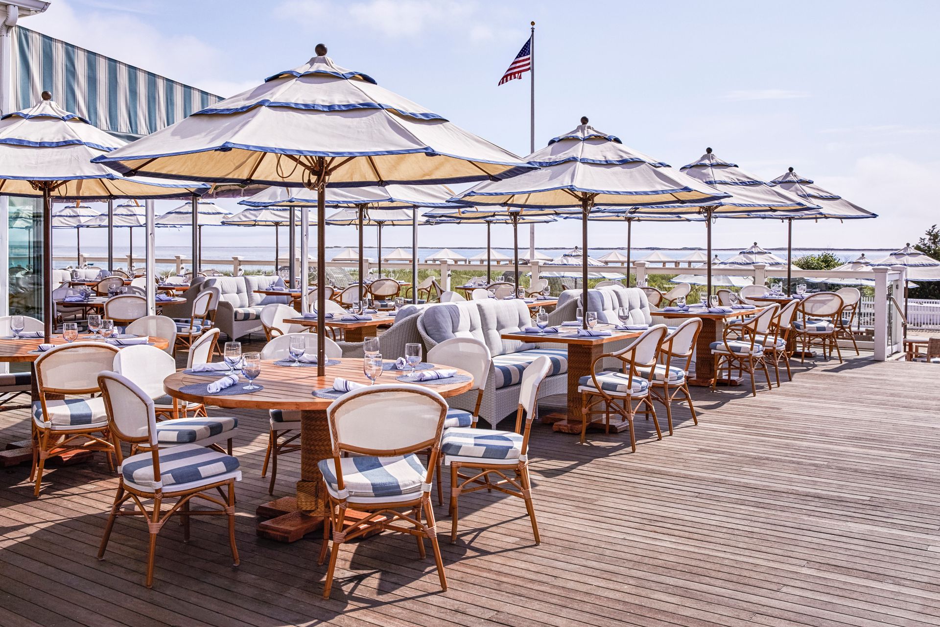 A beachfront outdoor restaurant with wooden tables, blue-striped chairs, and umbrellas, offering a scenic view by the water.