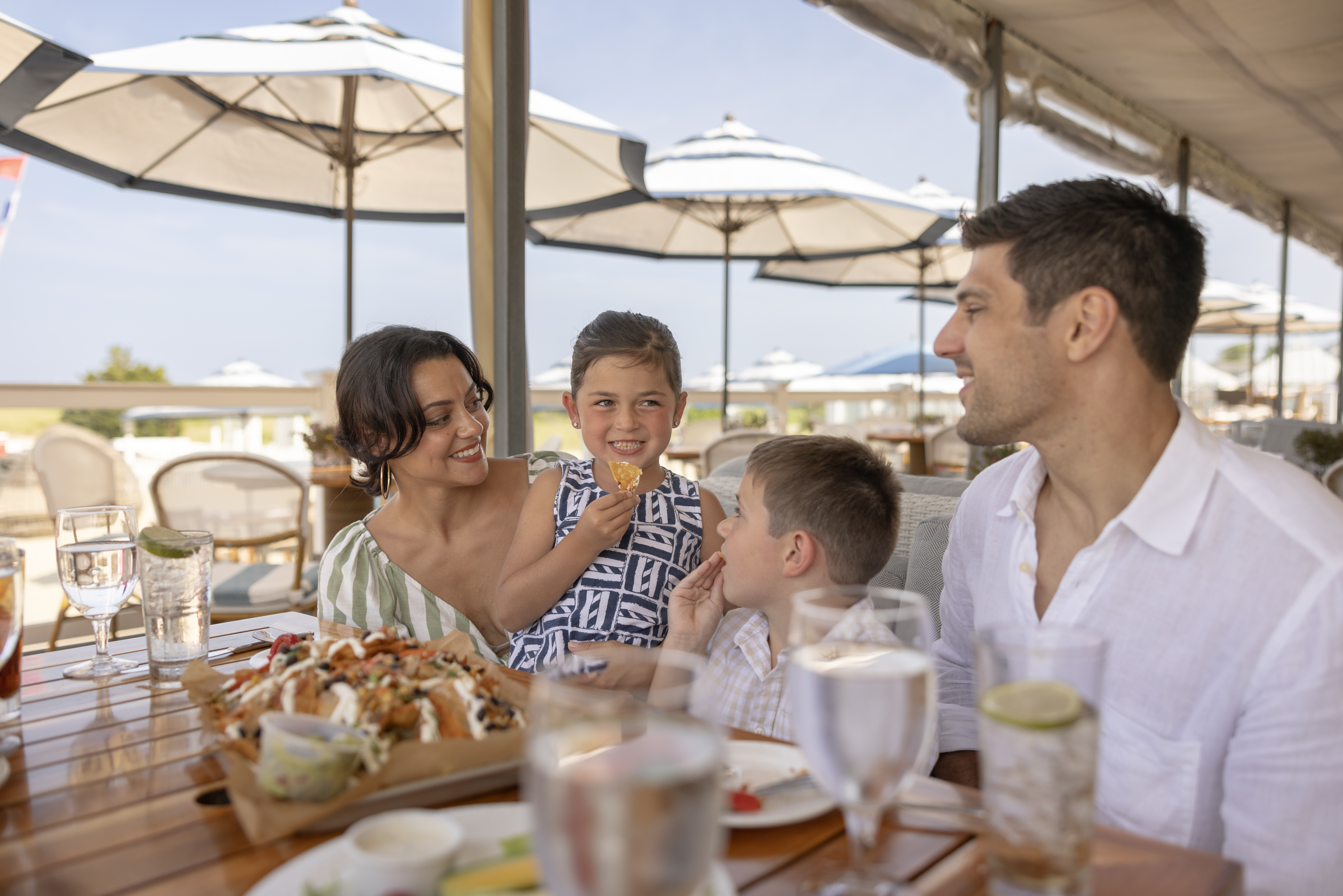 A family enjoys a meal together outdoors under umbrellas, with food and drinks on the table.