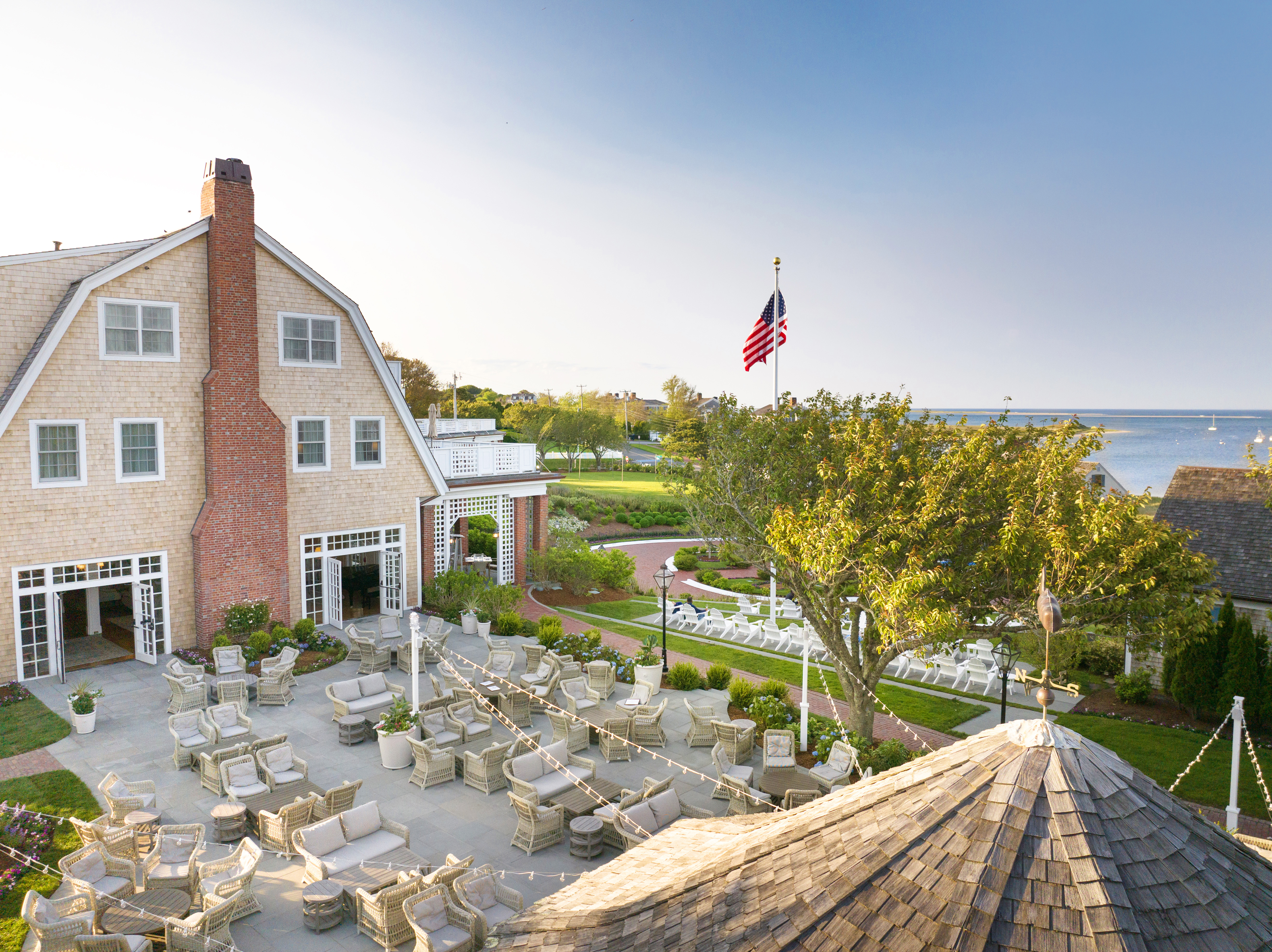 A scenic view of a coastal building with outdoor seating, an American flag, lush greenery, and a glimpse of the ocean in the background.