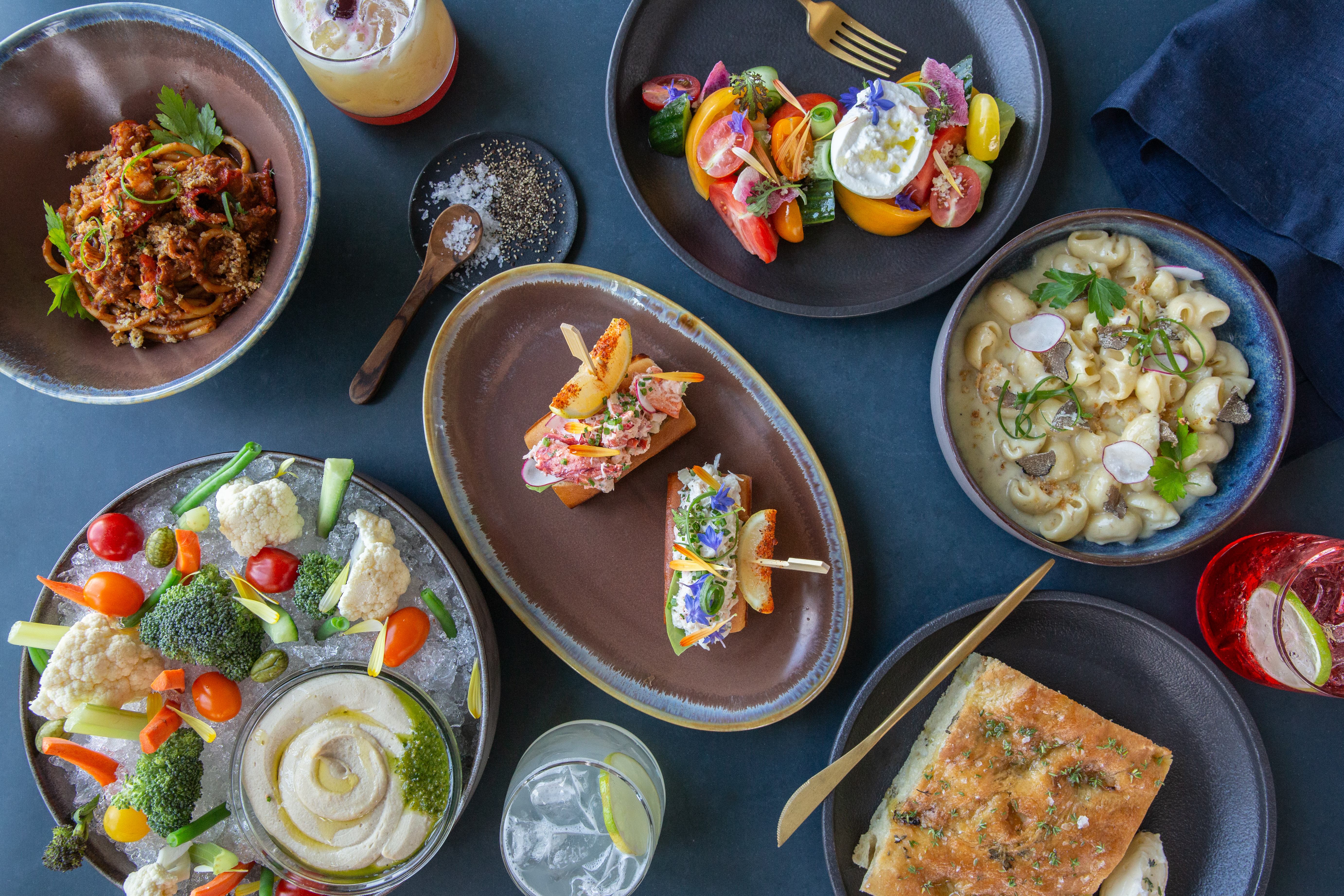 A variety of dishes including pasta, salad, dips with veggies, sliders, and drinks are artfully arranged on a table.