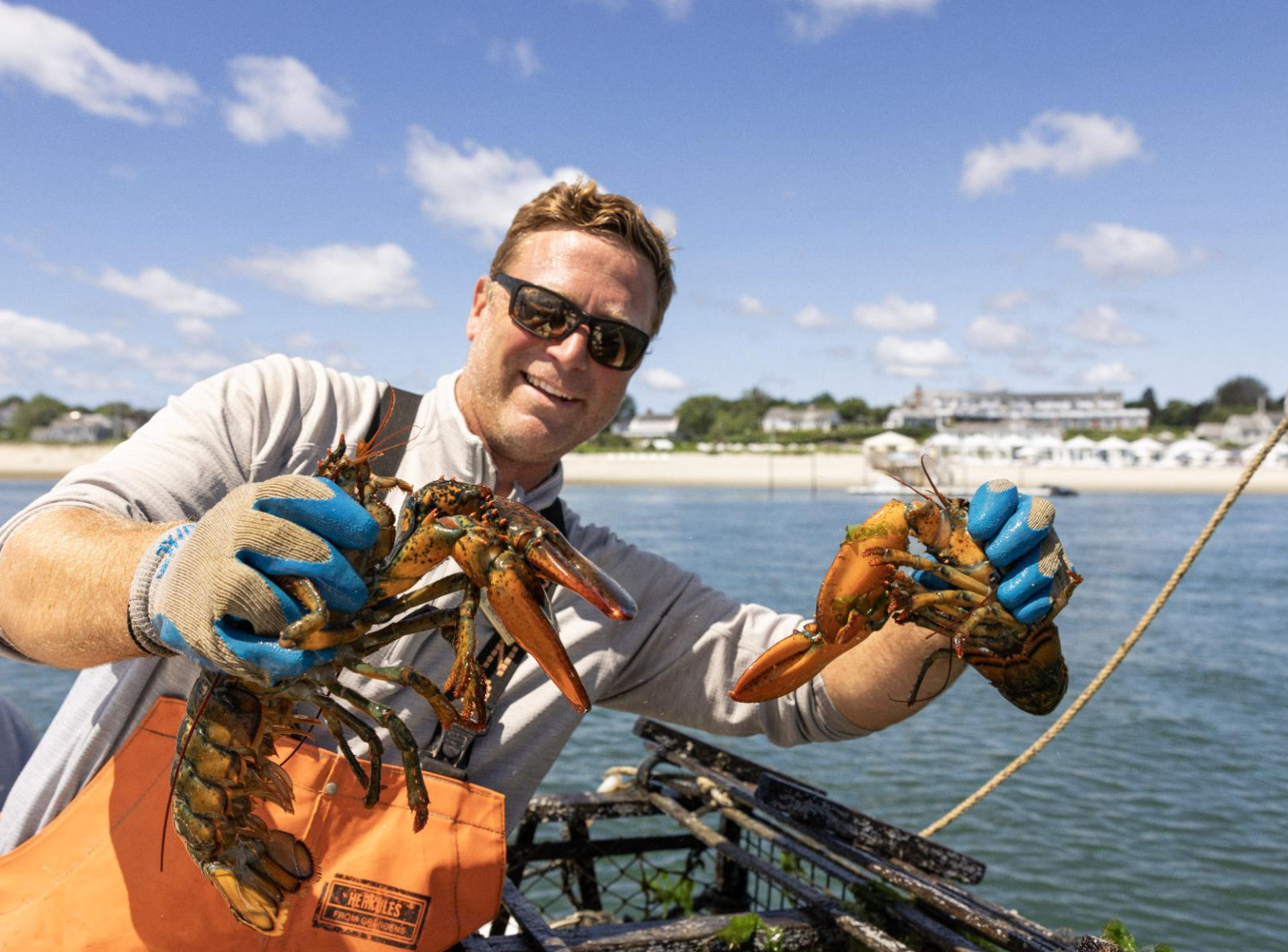 A person in sunglasses and gloves holds two lobsters near the sea, with a beach and buildings in the background.