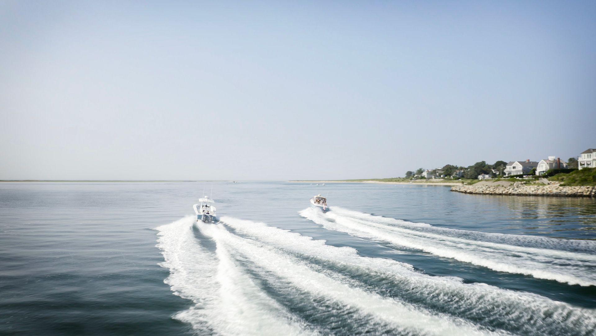 Two motorboats speed across the water leaving white wakes behind, with houses and greenery visible on the shoreline under a clear sky.