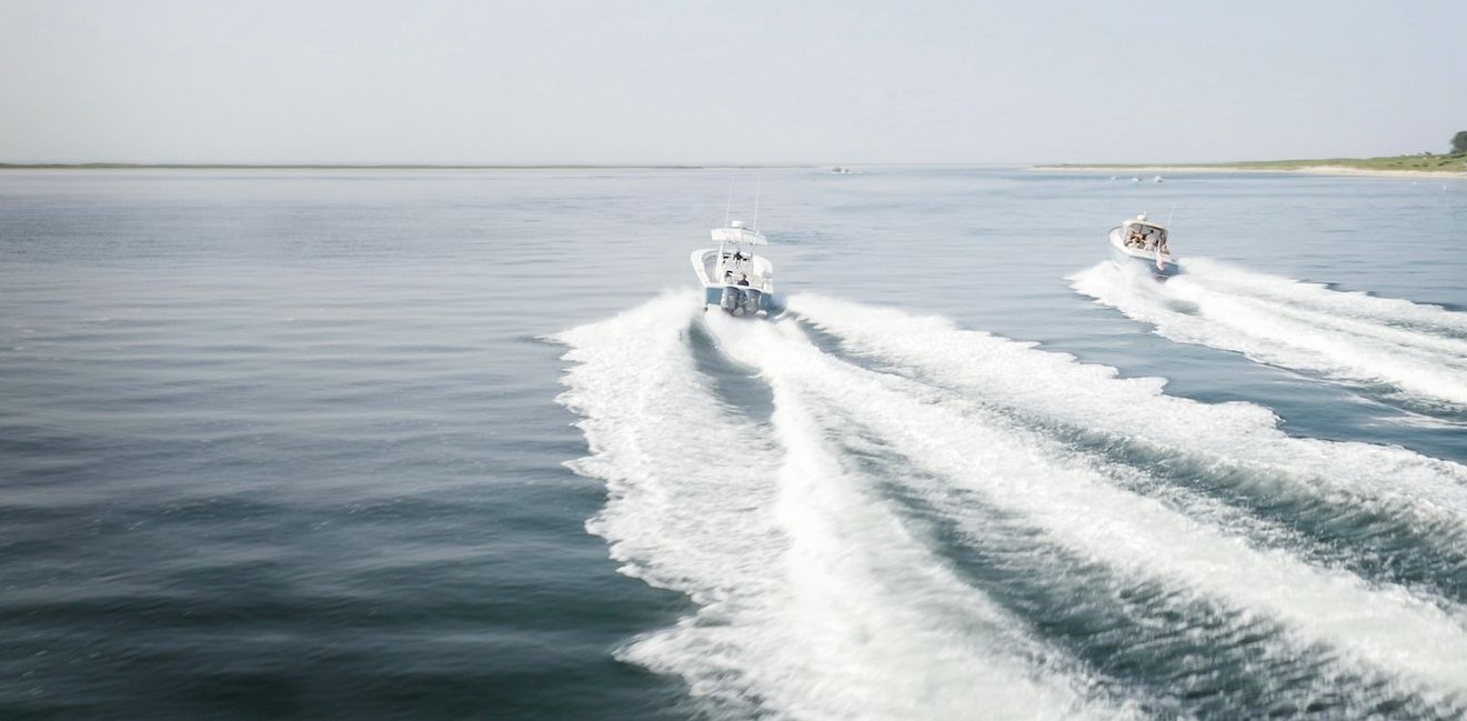 Two boats speed across calm waters leaving white wakes, with houses lining the distant shoreline under a clear blue sky.