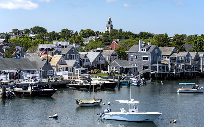 A scenic waterfront with boats, quaint houses, and a clock tower in the background, under a clear blue sky with some clouds showing.