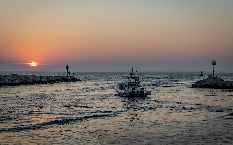 A boat sails between rocky jetties under a vibrant sunset, with calm waters reflecting the colorful sky.