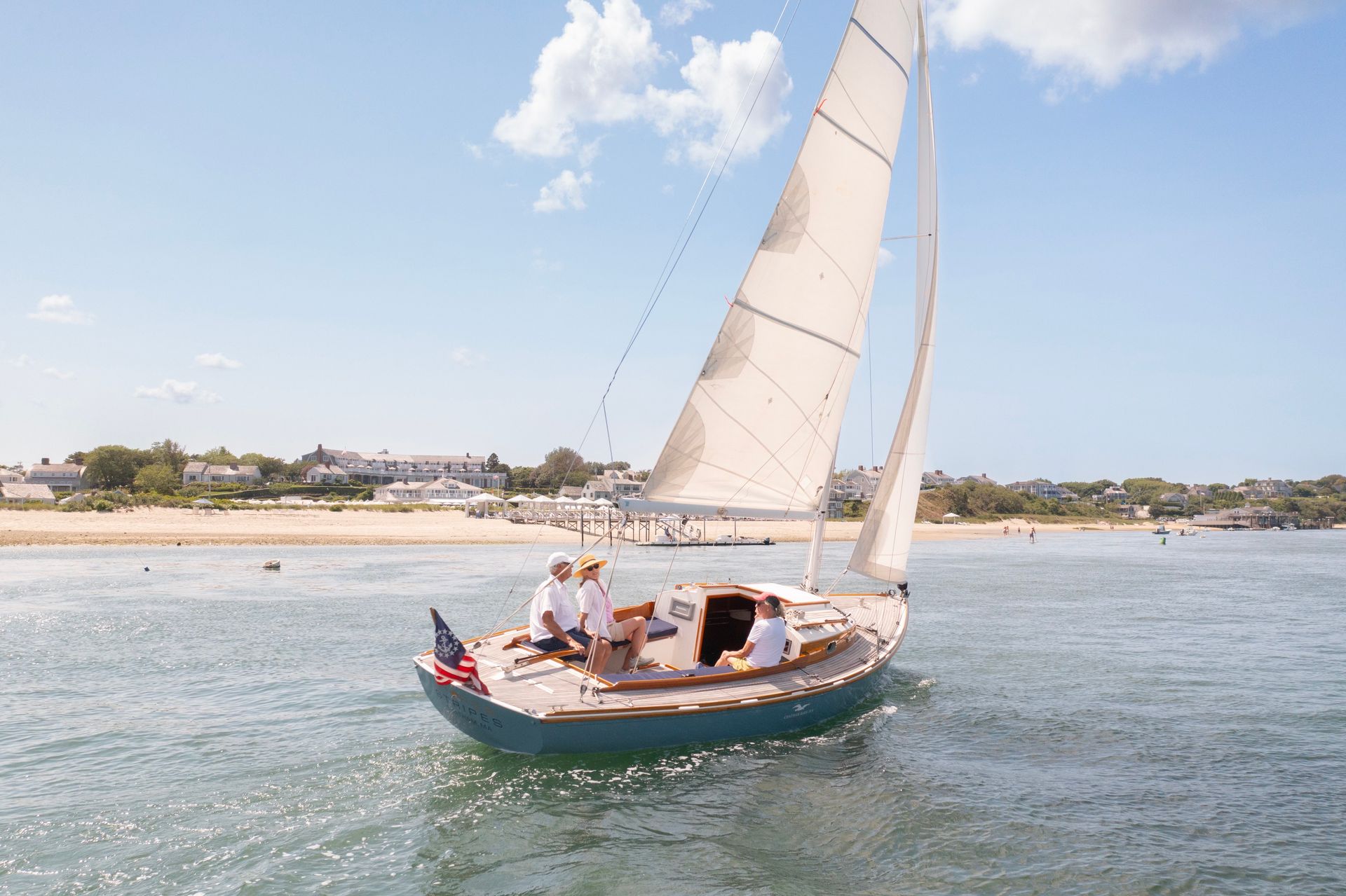 A sailboat with three people sails near a sandy shore under a clear blue sky, displaying an American flag at the stern.