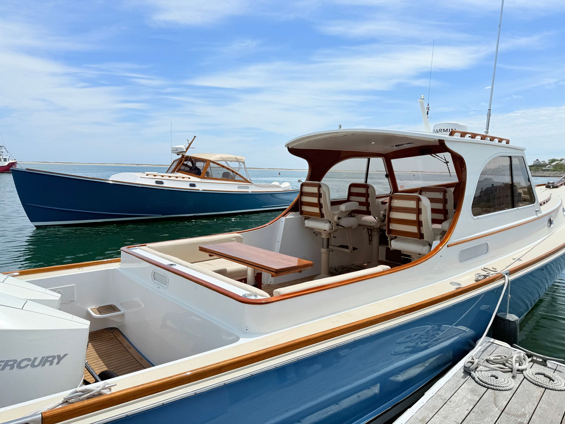 Two docked boats with blue hulls on a sunny day. The nearer boat has a wooden interior and seating with a covered helm area.