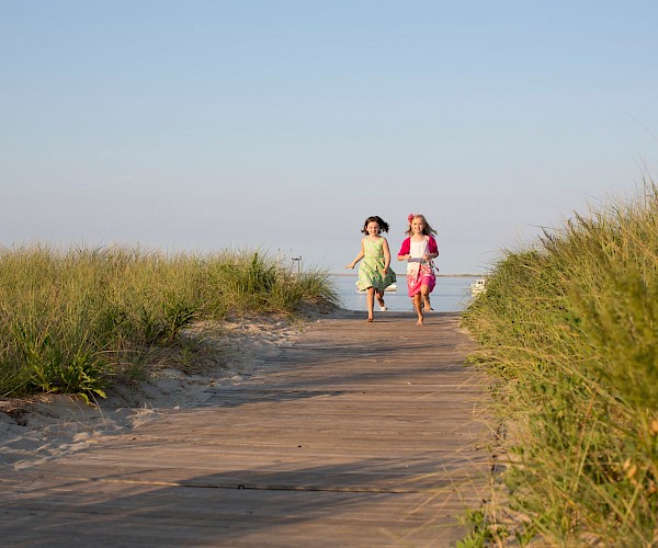 Two children run joyfully on a sandy path lined with grass, with the clear sky and sea in the background.