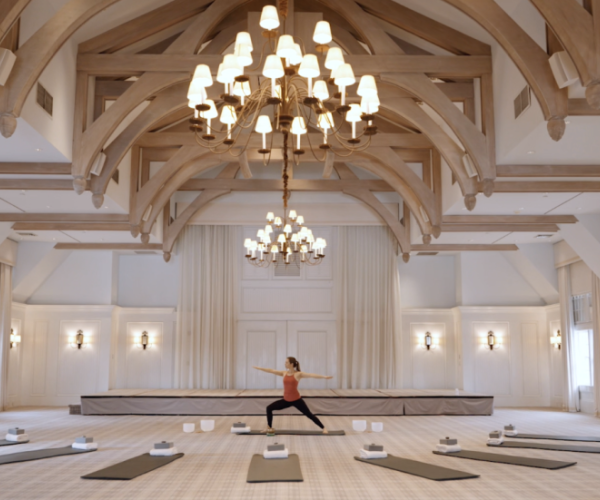 A person is doing yoga in an elegant room with chandeliers and yoga mats arranged in a semicircle on the floor.