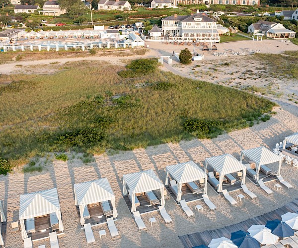 Aerial view of a beachside resort with cabanas, deck chairs, grassy areas, and adjacent buildings.