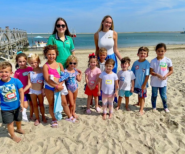 A group of children and two women pose on a sandy beach near a wooden dock, with the ocean and boats in the background.