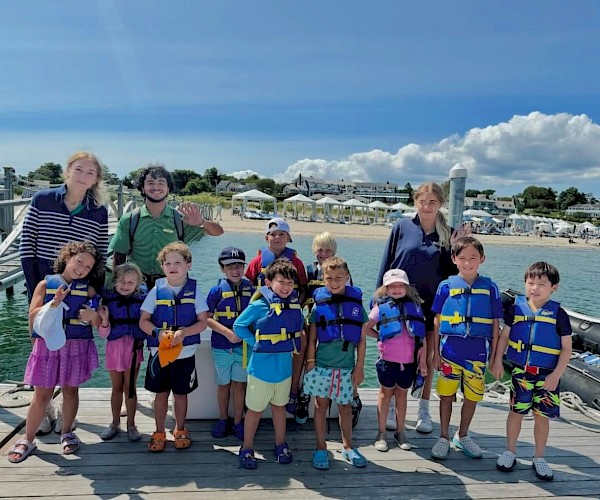A group of children in life vests with three adults pose on a dock by the water under a sunny blue sky.
