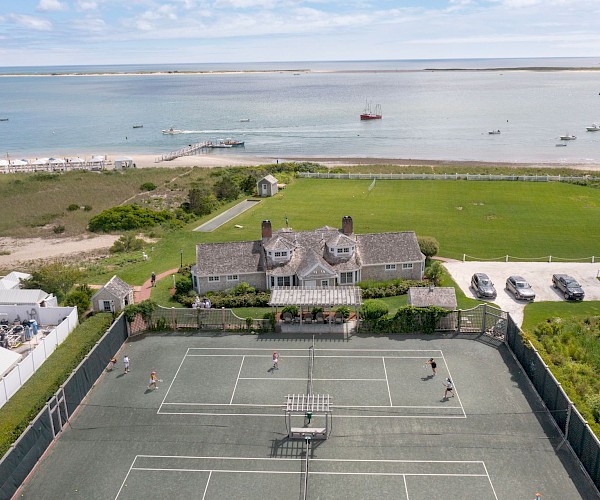 Aerial view of a seaside house with a tennis court and lawn, overlooking a beach with boats and a pier in the background.