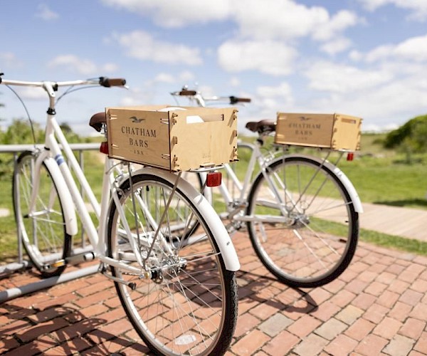Two white bicycles with "Chatham Bars Inn" boxes are parked on a brick path, with a person walking in the background.