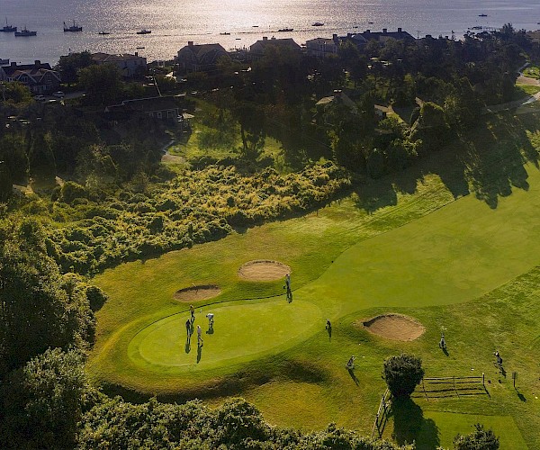 Aerial view of a golf course with players, near a coastline with boats and houses, surrounded by lush greenery.