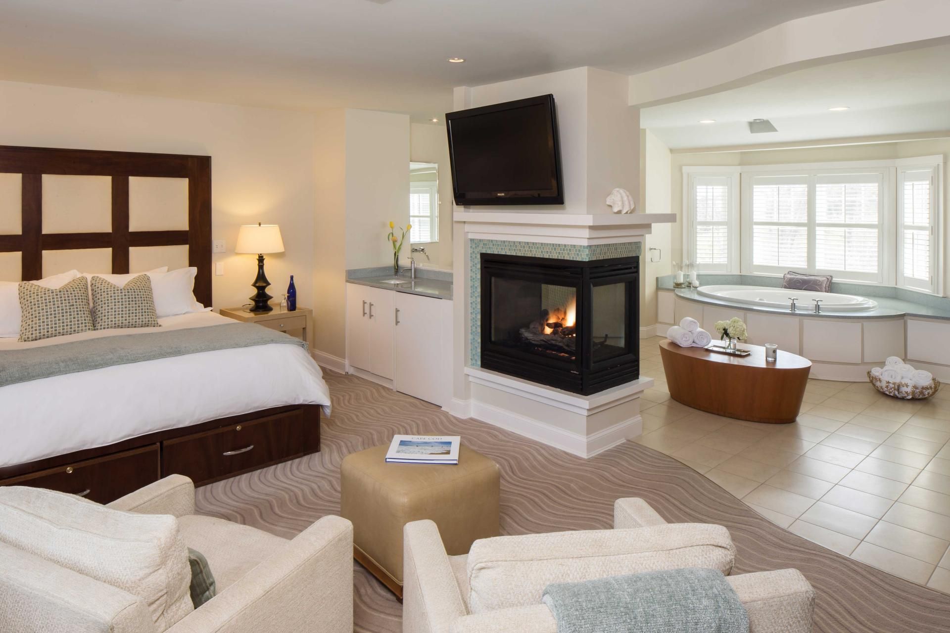 A cozy bedroom with a large bed, armchairs, a fireplace, and a bathtub. The decor is elegant and the room is well-lit.