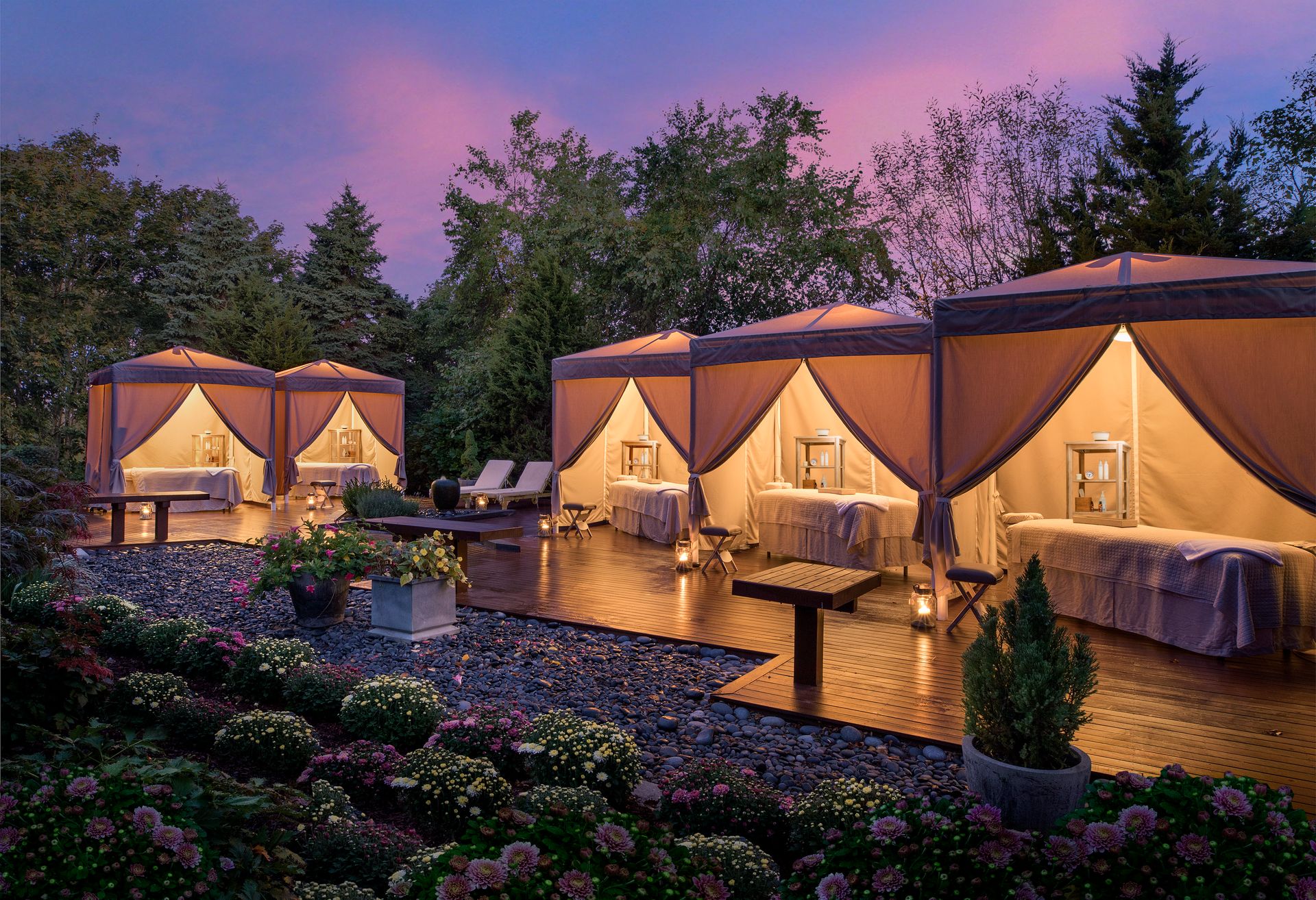 Outdoor massage cabanas with soft lighting, surrounded by gardens, create a serene atmosphere at sunset on a wooden deck.