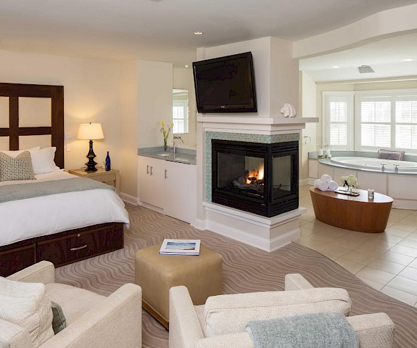A cozy bedroom with a fireplace, large bed, seating area, and a bathroom featuring a bathtub near bay windows, creating a luxurious feel.